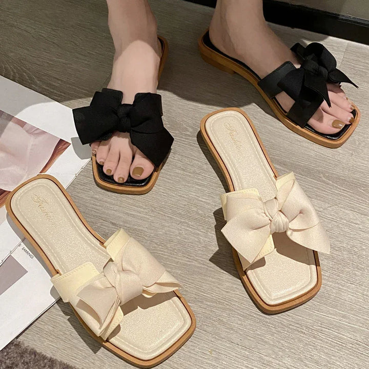 Pair of flat slide sandals with large bows, one worn in black and one cream on light wood floor