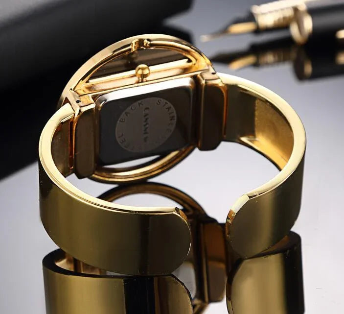 Close-up of a shiny gold cuff watch with a black stainless steel back on reflective surface