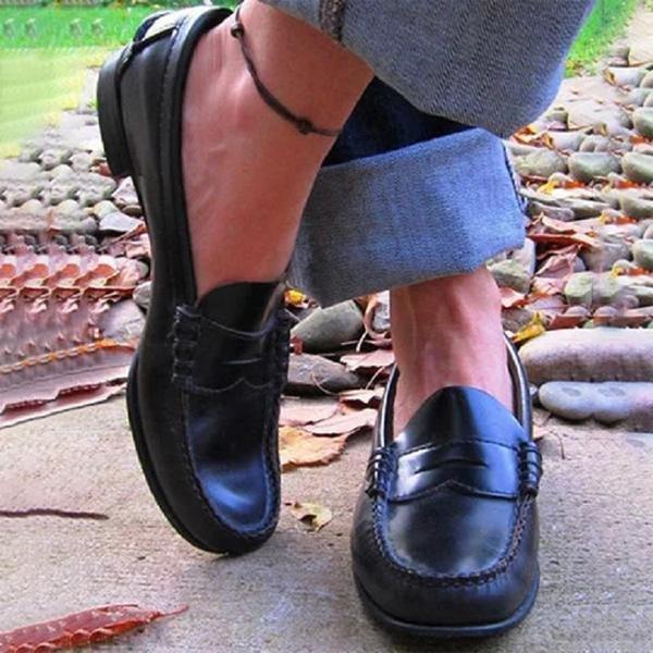 Close-up of black leather loafers worn with gray rolled-up pants on outdoor path with fallen leaves