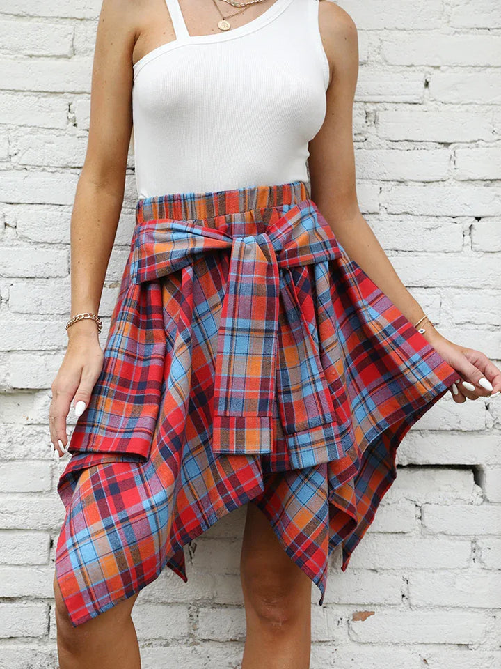 Woman wearing a red, blue, and orange plaid asymmetrical skirt with a tied waist bow and a white one-shoulder top