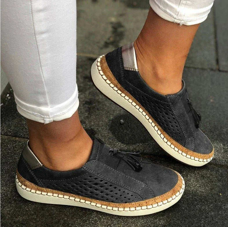 Black slip-on casual shoes with perforated sides, tassel detail, white sole, worn with white pants on pavement