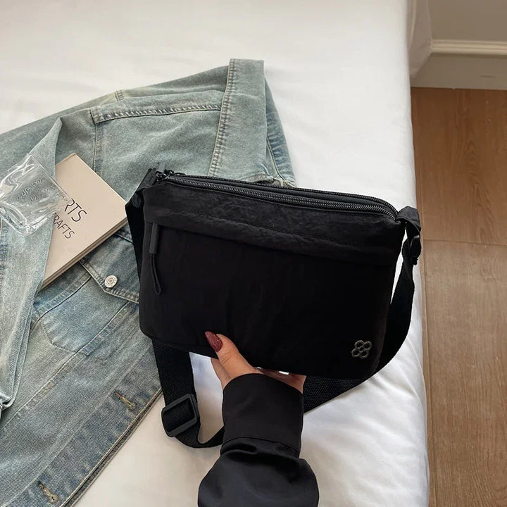 Black crossbody bag with zipper detail held over light denim jacket and book on white bed