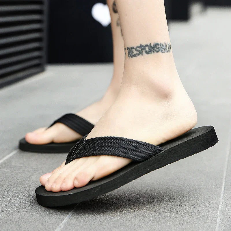 Close-up of feet wearing black fabric strap flip flops on gray tile floor with ankle tattoo