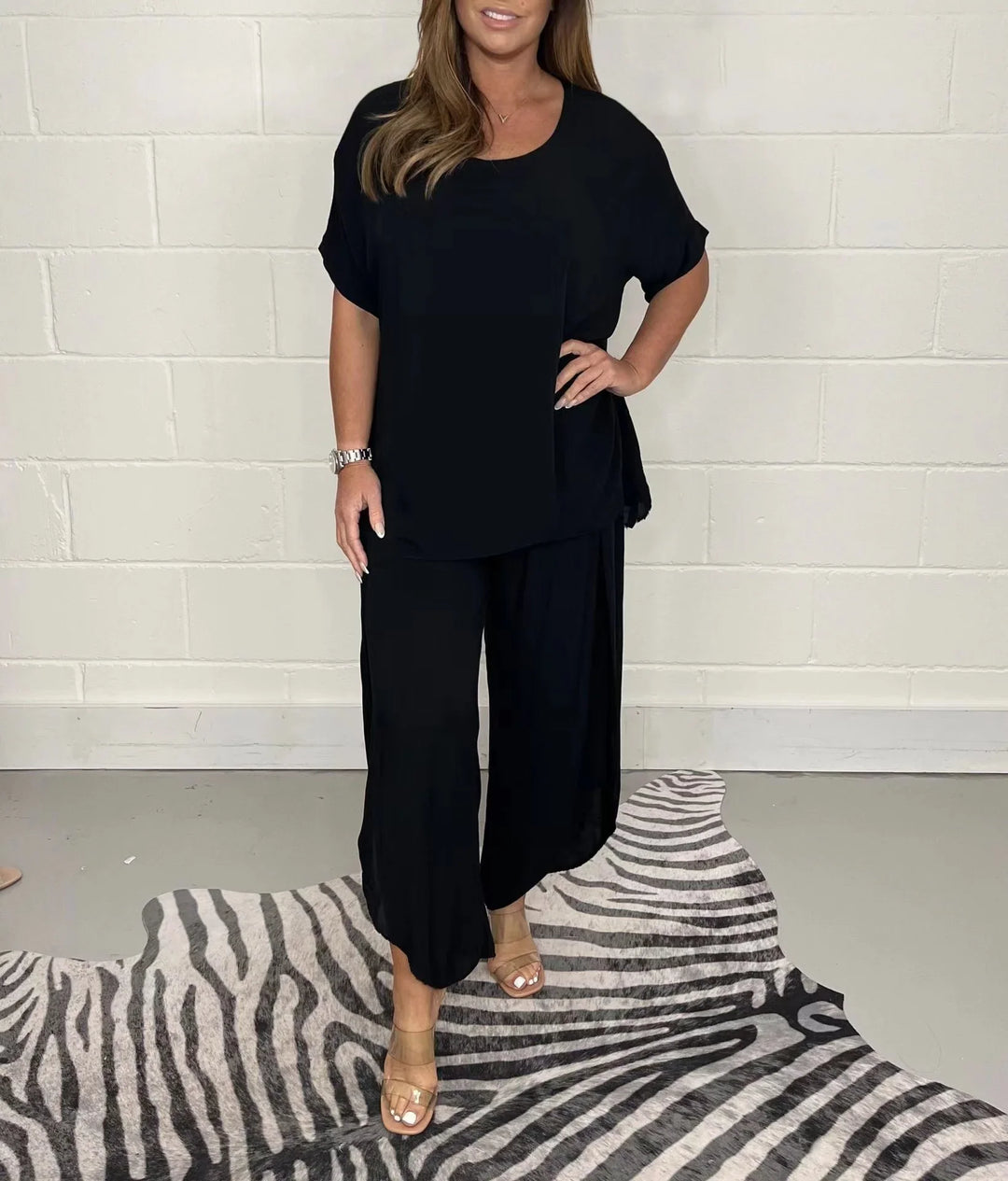 Woman wearing a black loose top and wide-leg pants standing on zebra pattern rug against a white brick wall