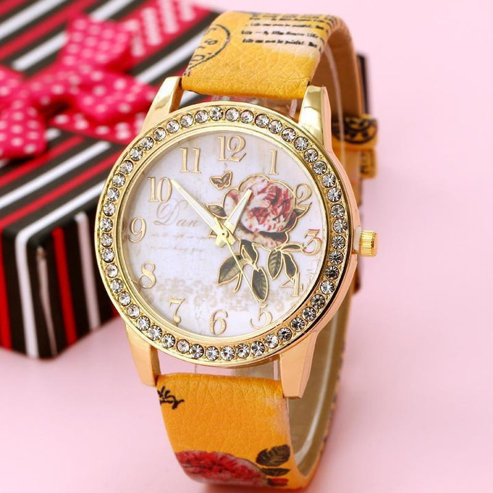 Gold wristwatch with crystal-studded bezel, floral dial design, and yellow leather strap on pink background