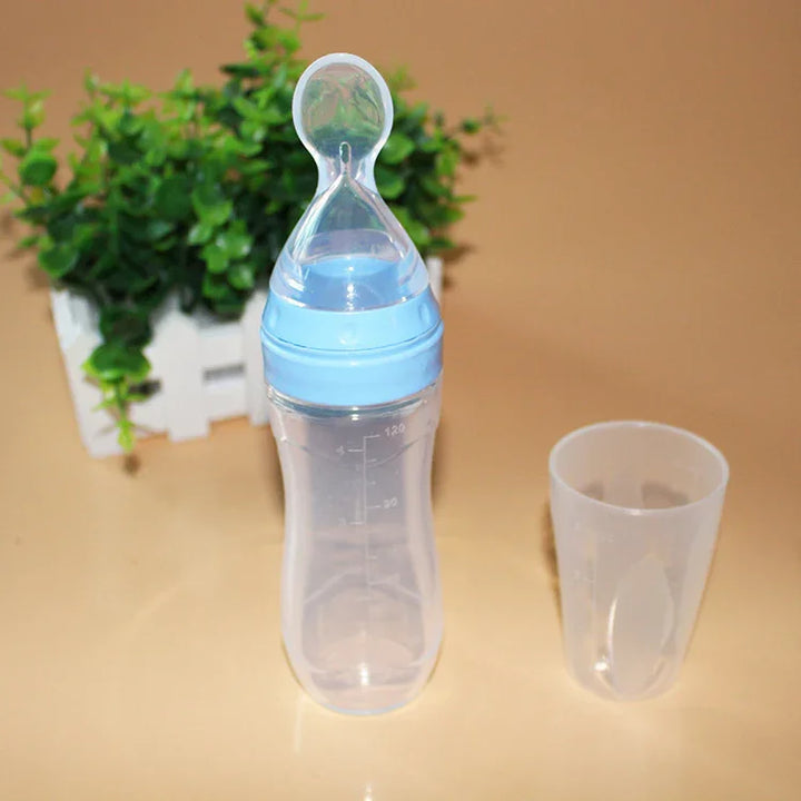 Baby feeding bottle with blue cap and transparent measuring cup on beige surface with green plant background
