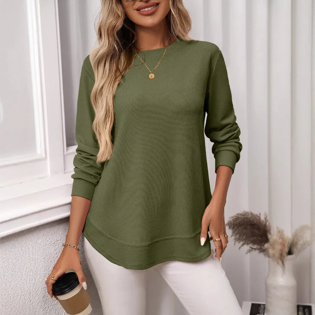 Woman wearing olive green long-sleeve knit sweater with white pants holding coffee cup indoors