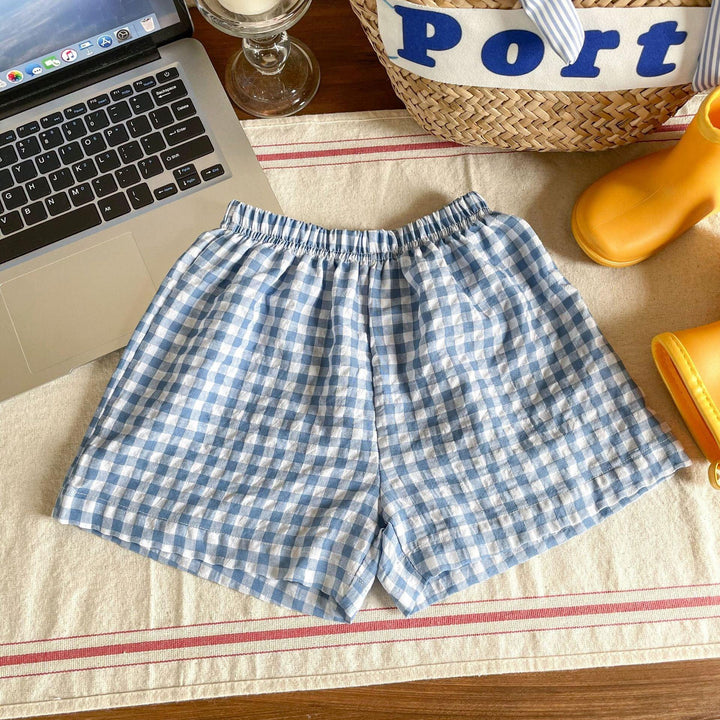 Blue and white checkered cotton shorts on table with laptop, glass, basket, and yellow boots