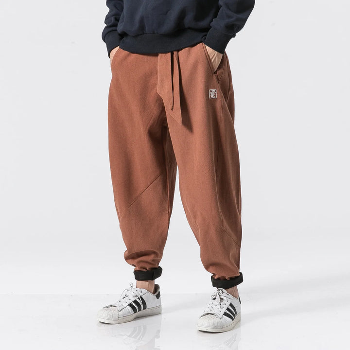 Model wearing rust brown relaxed fit pants with a drawstring waist and white sneakers with black stripes