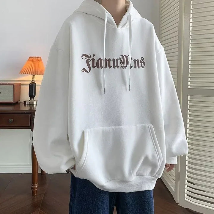 White oversized hoodie with black gothic text 'JianuMns' worn indoors by person near wooden desk and lamp