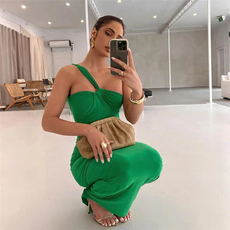 Woman in green one-shoulder dress taking mirror selfie, holding woven clutch and wearing gold accessories