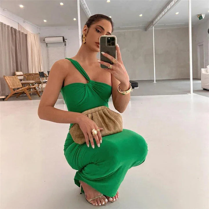 Woman in green one-shoulder dress taking mirror selfie, holding woven clutch and wearing gold accessories