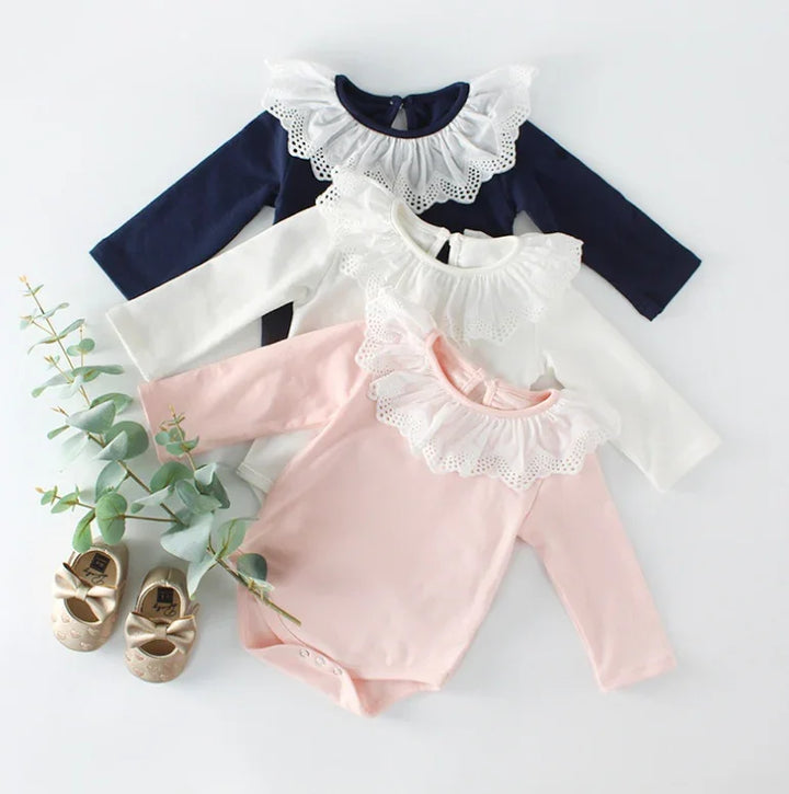 Three long-sleeve baby bodysuits in navy, white, and pink with white lace collars, paired with beige baby shoes
