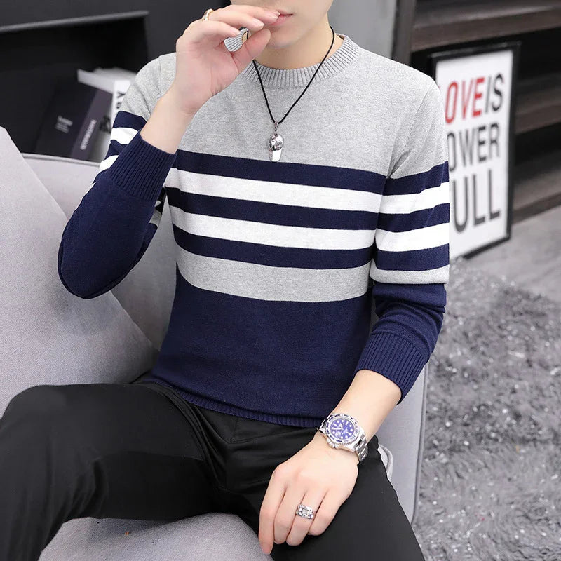 Man wearing gray and navy striped sweater with black pants, silver watch, and pendant necklace sitting on gray sofa