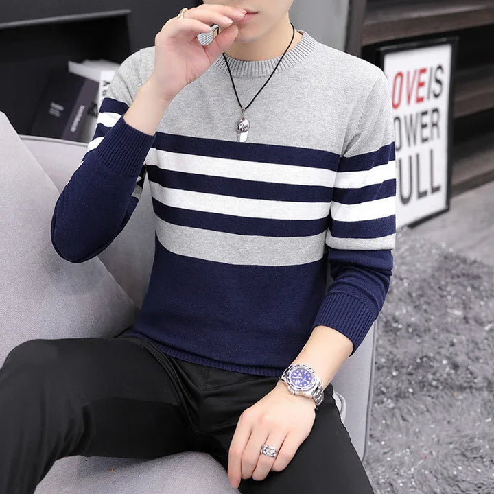Man wearing gray and navy striped sweater with black pants, silver watch, and pendant necklace sitting on gray sofa