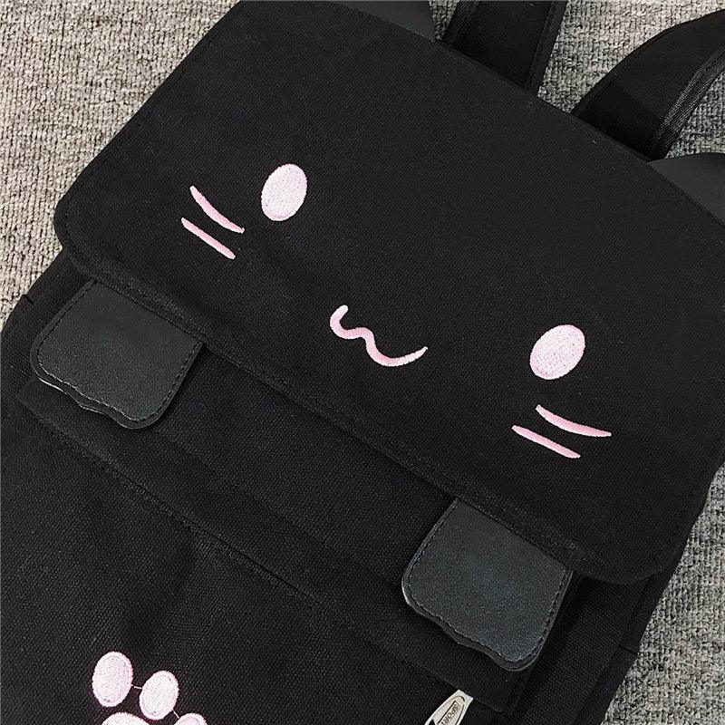 Black canvas backpack with cute pink cat face embroidery and faux leather straps on gray carpet