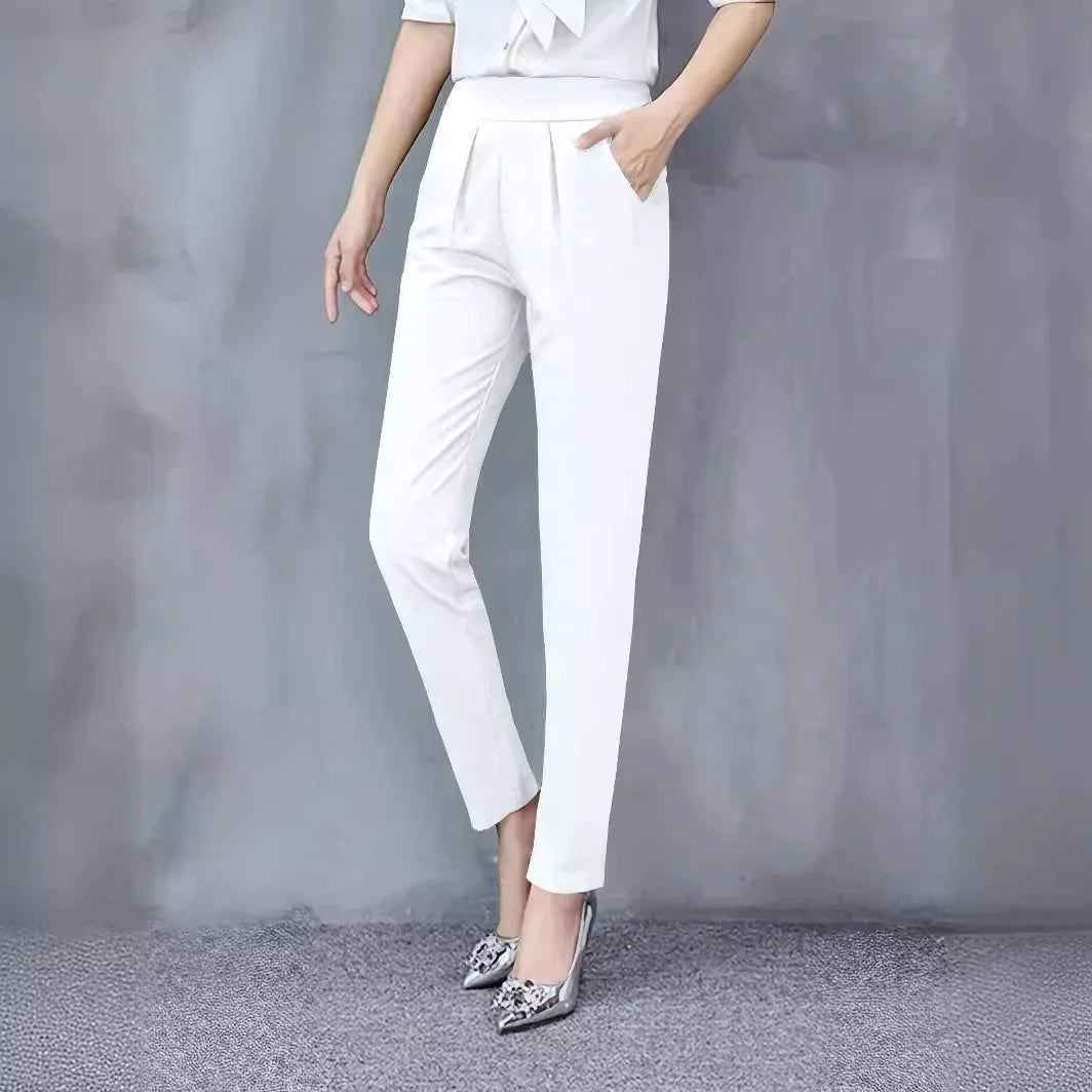 Woman wearing white tapered pants and silver pointed high heels standing against grey background