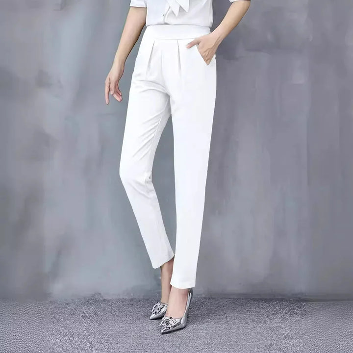Woman wearing white tapered pants and silver pointed high heels standing against grey background