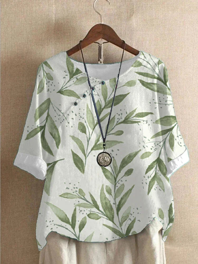 White short-sleeve blouse with green leafy print and button details, paired with a necklace on wooden hanger