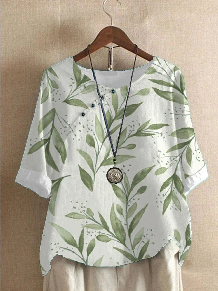 White short-sleeve blouse with green leafy print and button details, paired with a necklace on wooden hanger