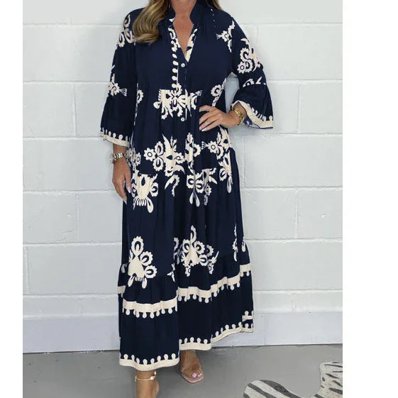 Woman wearing navy blue maxi dress with white floral print and tassel hem, standing against white brick wall
