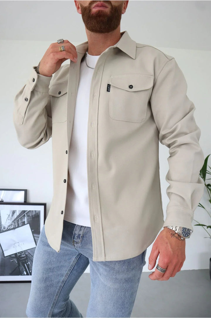 Man wearing beige button-up shirt with chest pockets over white t-shirt and light blue jeans in modern room