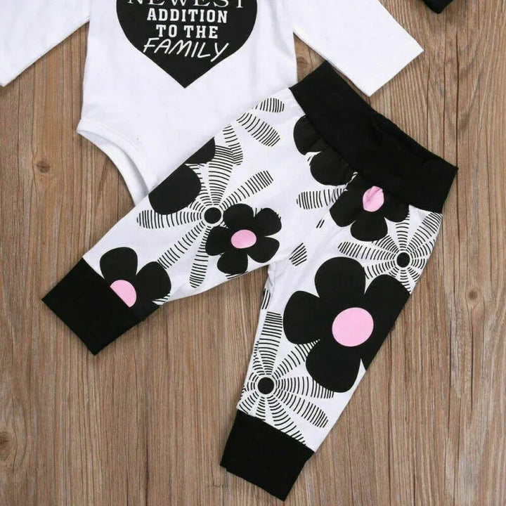 Baby outfit with white onesie reading newest addition to the family and black floral print pants with pink centers