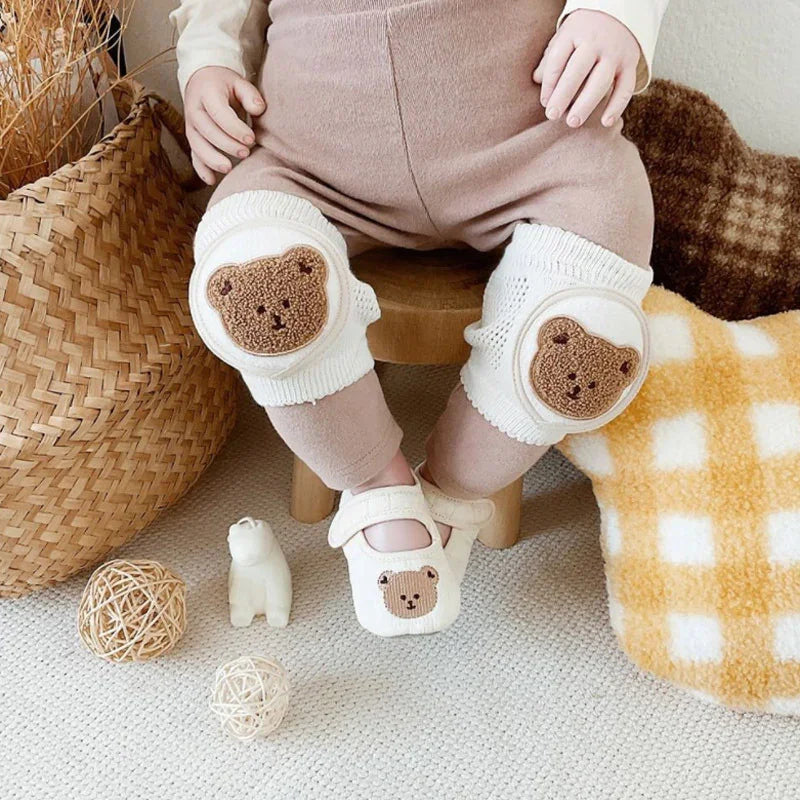 Baby wearing beige pants with white knee pads and shoes featuring cute teddy bear faces, seated on wooden stool