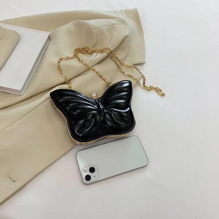Black butterfly-shaped handbag with gold chain on beige coat next to white smartphone on white surface