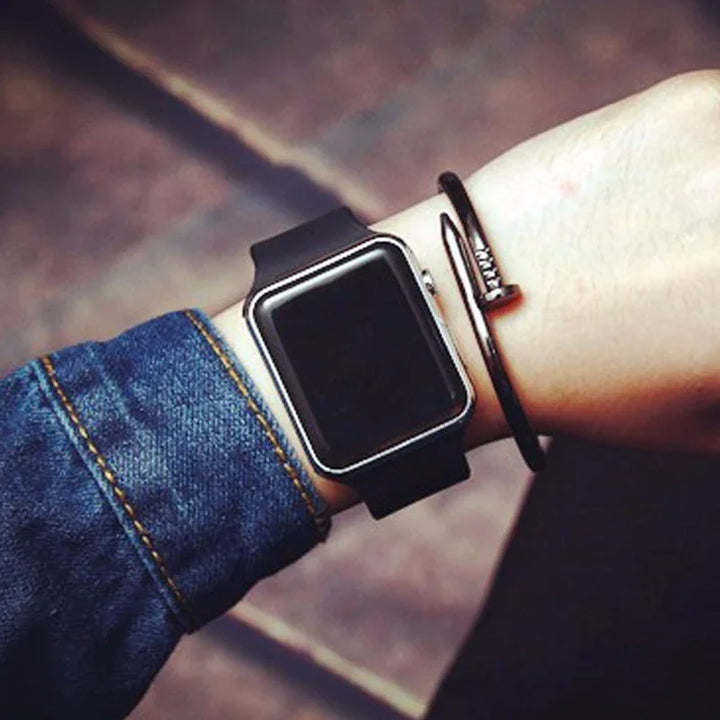 Wrist wearing a black smart watch with a rectangular screen and a black nail-shaped bracelet, dressed in a denim jacket sleeve