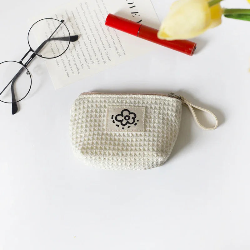 Textured cream pouch with flower patch, eyeglasses, red lipstick, paper, and yellow flower on white surface