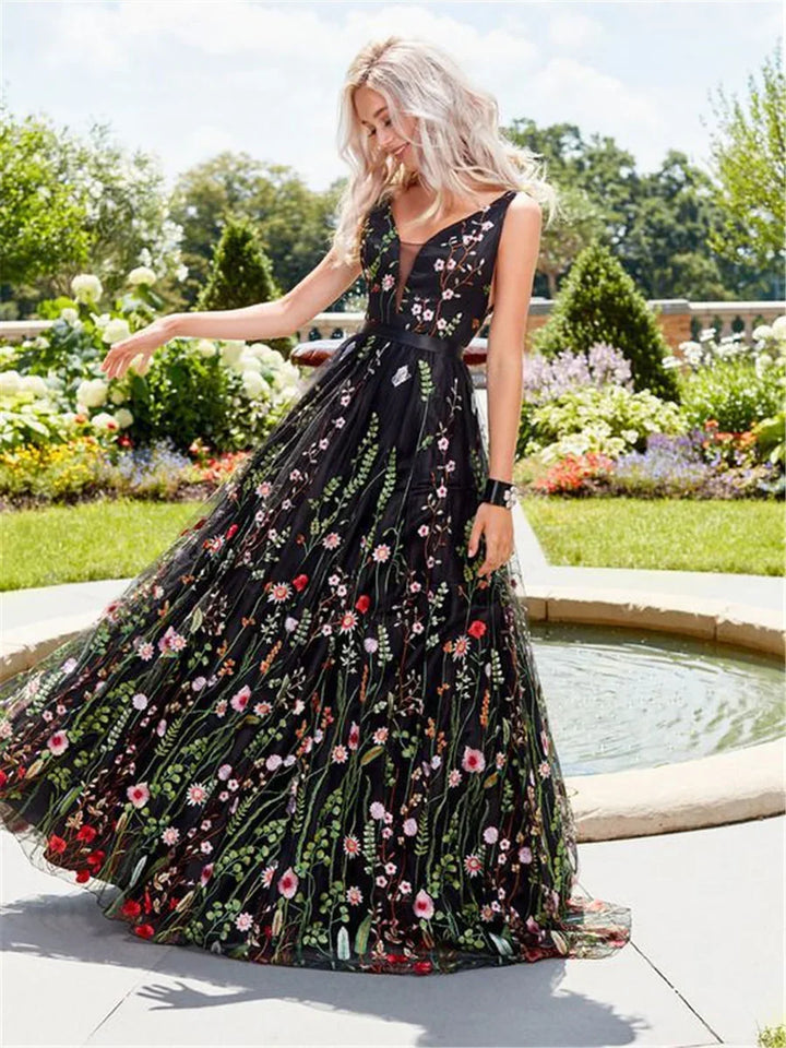 Blonde woman in black floral embroidered gown posing outdoors near garden fountain on sunny day