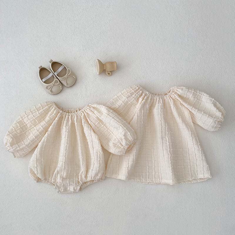 Cream textured baby outfit set with puff sleeves, matching dress, beige baby shoes, and wooden rattle on white background