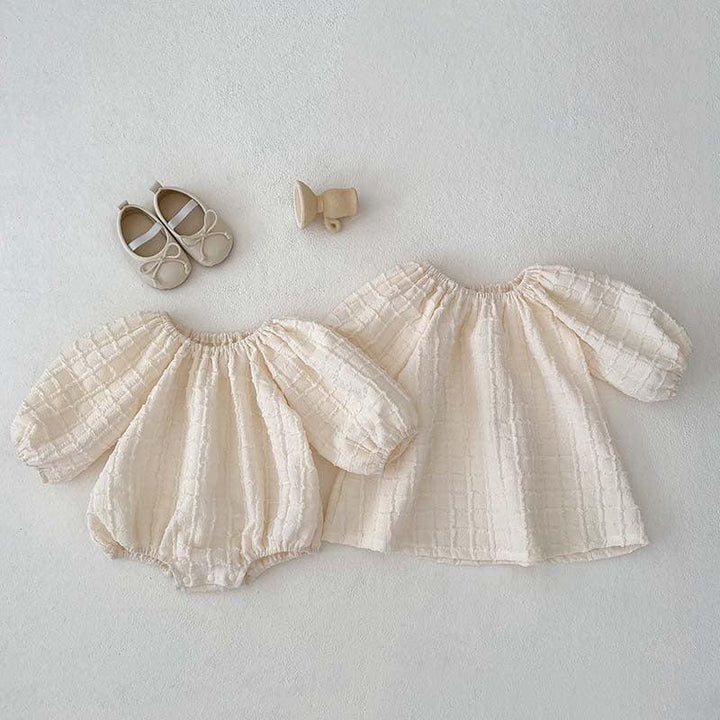 Cream textured baby outfit set with puff sleeves, matching dress, beige baby shoes, and wooden rattle on white background