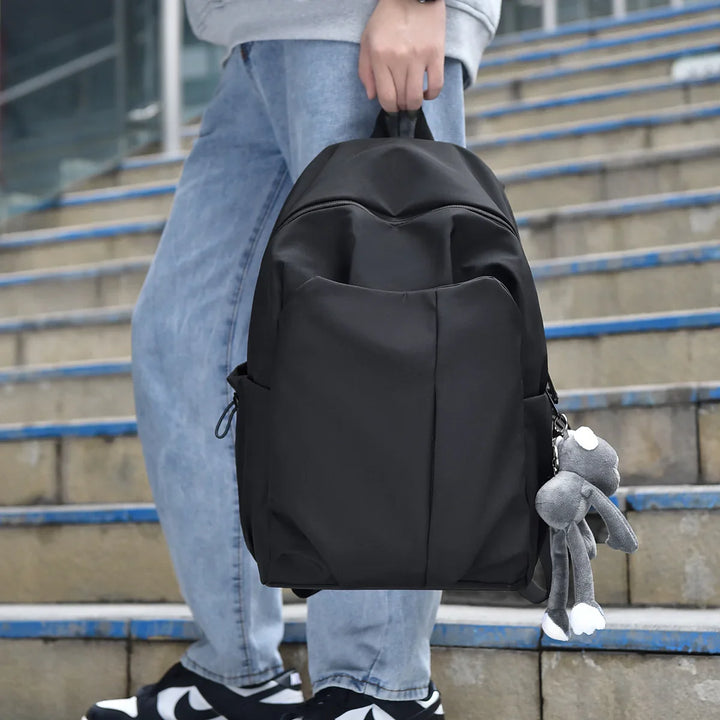 Person holding black backpack with gray plush keychain on outdoor stairs, wearing casual jeans and sneakers