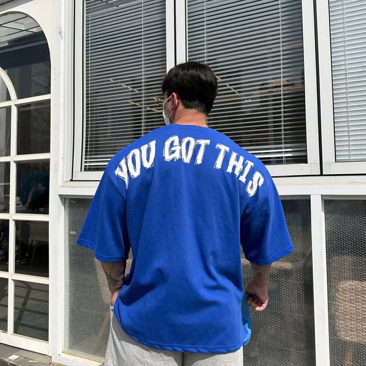 Man wearing blue oversized t-shirt with white text You Got This standing outdoors by window blinds