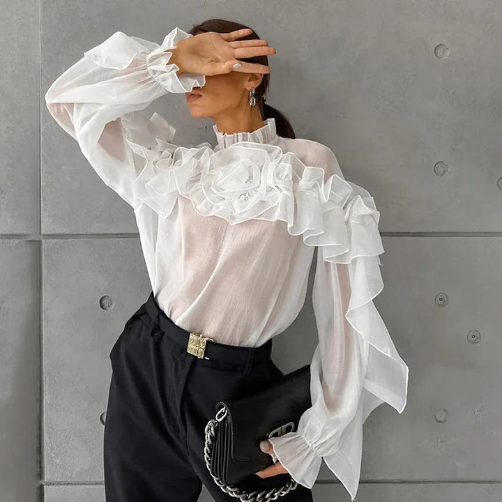 Woman wearing a sheer white blouse with ruffle details and black pants, holding a black purse with chain