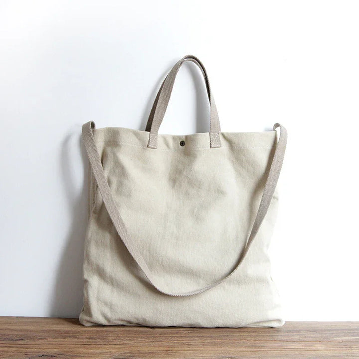 Beige canvas tote bag with short handles and long shoulder strap on wooden surface