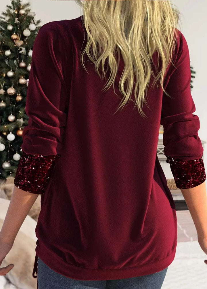 Woman wearing burgundy long sleeve top with sequined cuffs standing in cozy living room with decorated Christmas tree