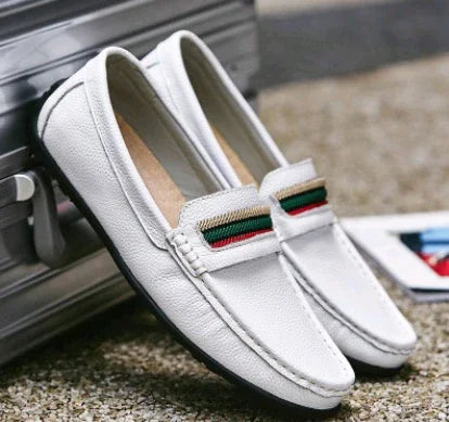 White leather loafers with green and red striped detail, placed against a silver suitcase on gravel