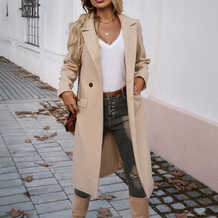 Woman wearing beige long coat with white V-neck top, gray distressed jeans, and beige boots outdoors