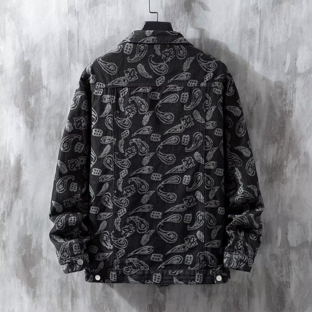Black denim jacket with grey paisley pattern hanging on a hanger against textured grey wall