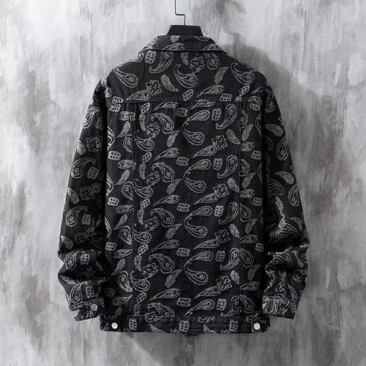 Black denim jacket with grey paisley pattern hanging on a hanger against textured grey wall