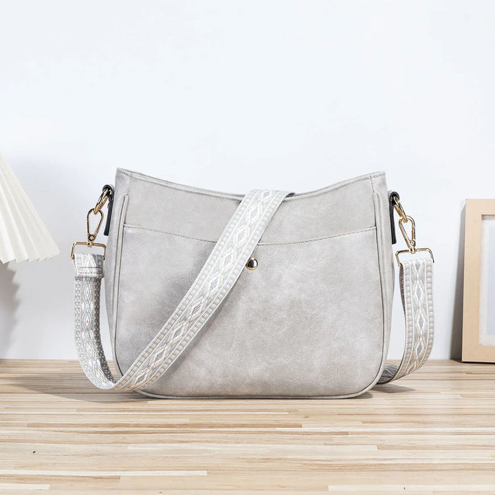Light gray leather crossbody handbag with patterned wide shoulder strap on wooden floor