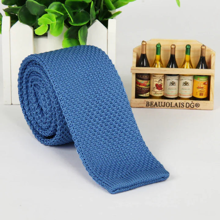 Rolled blue knitted men's tie displayed on white surface with green plant and miniature wine bottle crate