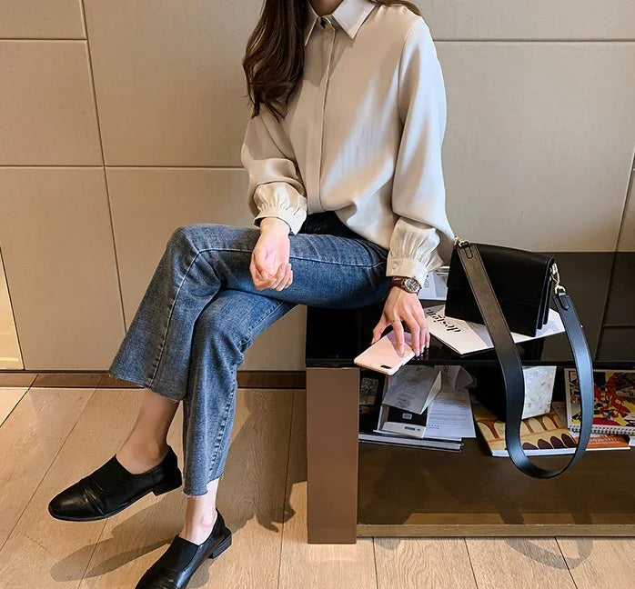 Woman wearing beige blouse and blue jeans sitting on a bench holding a smartphone with black leather handbag beside her