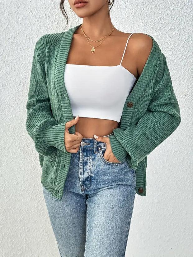 Woman wearing a green oversized knit cardigan over a white cropped camisole and high-waisted blue jeans against a textured white wall
