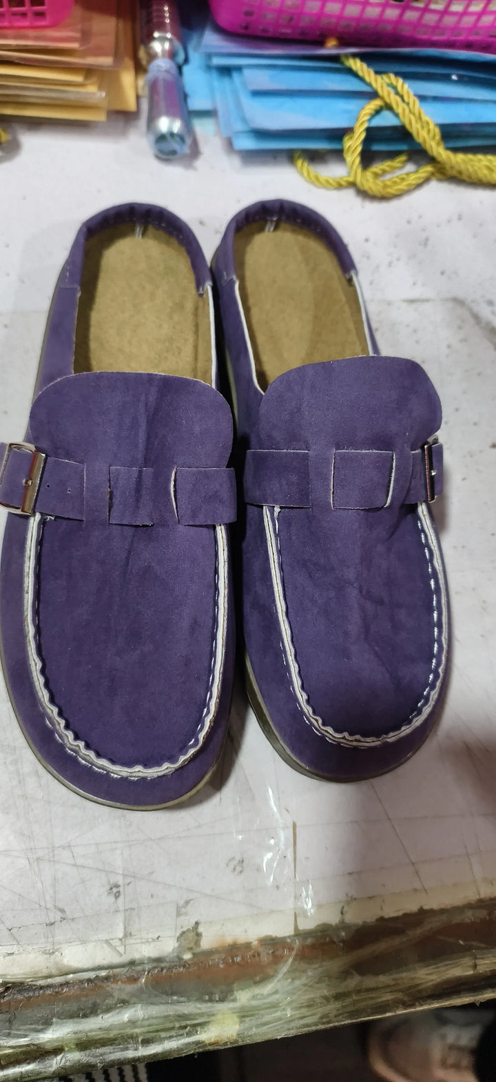 Pair of purple suede loafers with white stitching and buckle detail on a cluttered desk