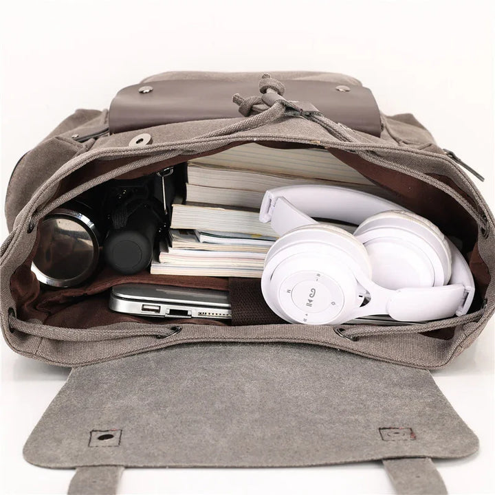 Open gray canvas backpack filled with white headphones, stacked books, laptop, and water bottle on white background