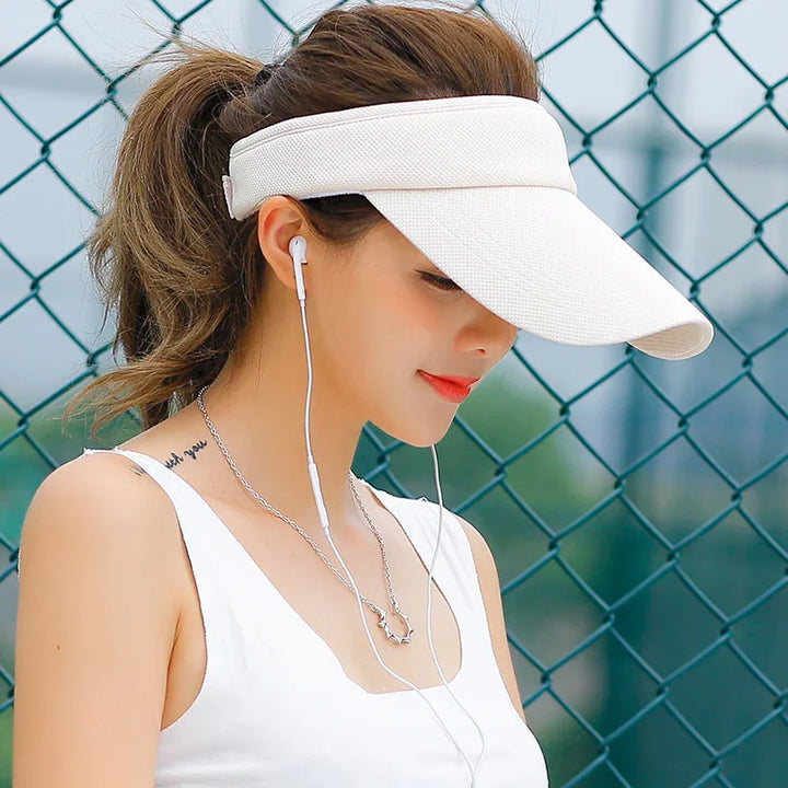 Young woman wearing a white sports visor, white tank top, and earphones, standing by a green chain-link fence